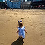 child, toddler, beach, sand, shadow, blue_bow, dress, barefoot, walking, sunny, outdoor, daytime, people, colorful_houses, background, summer, vacation, shore, coast, leisure