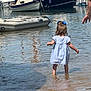 child, toddler, water, marina, boats, dress, bow, barefoot, reflection, summer, outdoor, exploring, shore, hand, person, small_boat, inflatable_boat, ripples, daytime, curious