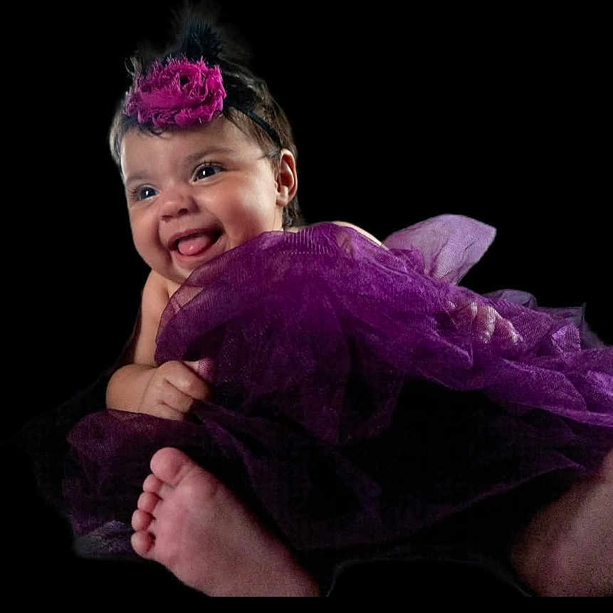 Nouva is registered to the contest to win money with this photo: baby, black_background, cheerful, child, closeup, clothing, cute, cute_expression, face, flower, foot, happy, headband, infant, person, portrait, purple_tutu, smiling, studio, tulle
