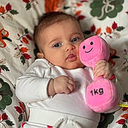 Emma is registered to the contest to win money with this photo: baby, blanket, child, close_up, cute, dumbbell, face, floral_pattern, hand, happy, indoor, infant, lying_down, person, pink, plush_toy, smile, soft_toy, toy, white_clothing
