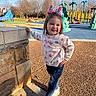 child, girl, playground, outdoor, smiling, casual_clothing, jeans, sneakers, bunny_ears, spring, sunlight, shadow, concrete, stone_wall, trees, play_equipment, happy, cute, posing, daytime