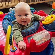 Steven joined the competition — help win amazing prizes! baby, infant, smiling, face, chubby_cheeks, activity_center, toy, steering_wheel, play_seat, colorful, red_plastic, blue_cushion, crib, indoor, hand, eyes, clothing, closeup, happy, floor
