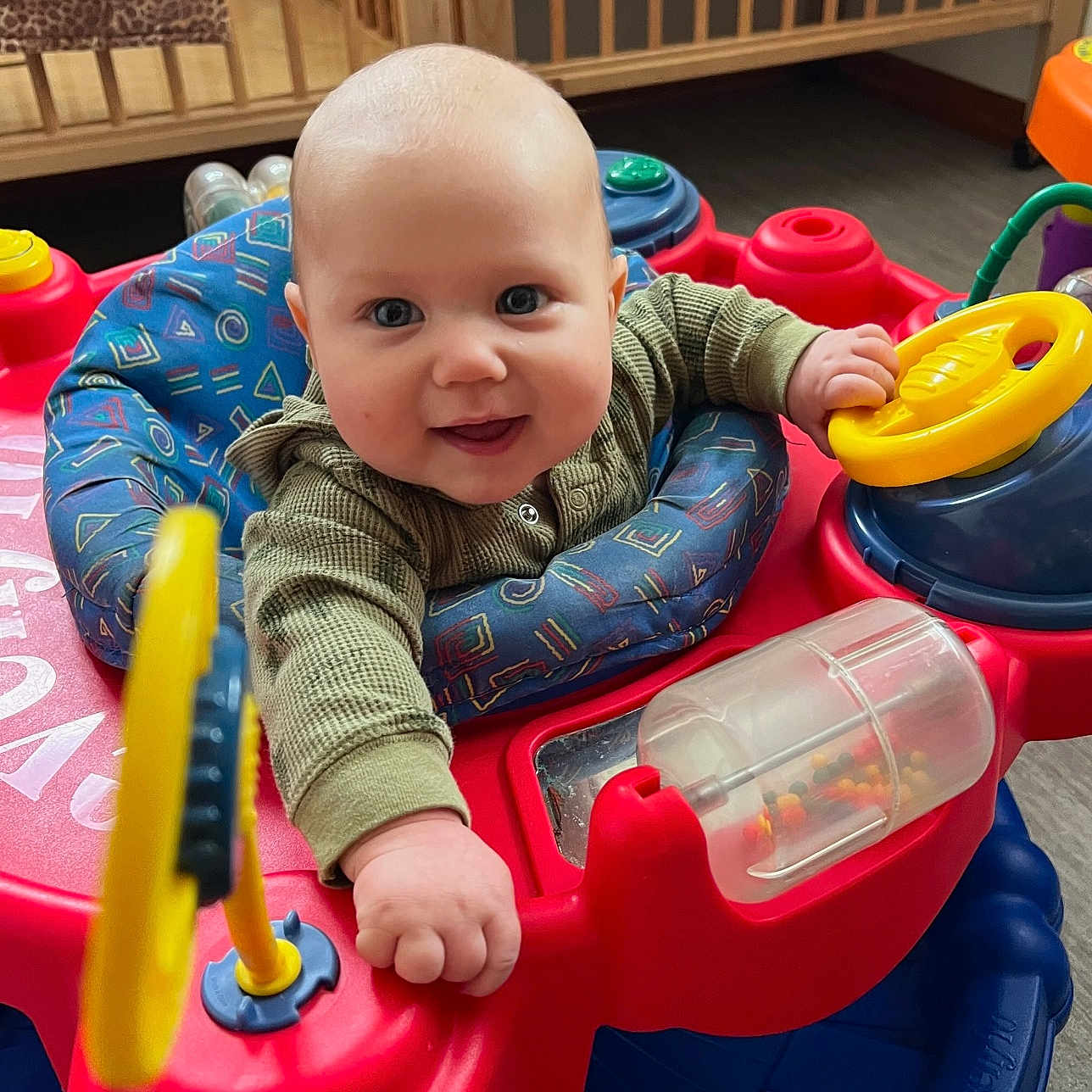Steven joined the competition — help win amazing prizes! activity_center, baby, blue_cushion, chubby_cheeks, closeup, clothing, colorful, crib, eyes, face, floor, hand, happy, indoor, infant, play_seat, red_plastic, smiling, steering_wheel, toy