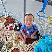 Caius is registered to the contest to win money with this photo: baby, child, playpen, quilt, plush_toy, blue_toy, smiling, indoor, floor, happy, curly_hair, clothing, short_sleeve, toy, colorful, patterned, sitting, person, face, cute
