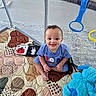 baby, child, playpen, quilt, plush_toy, blue_toy, smiling, indoor, floor, happy, curly_hair, clothing, short_sleeve, toy, colorful, patterned, sitting, person, face, cute