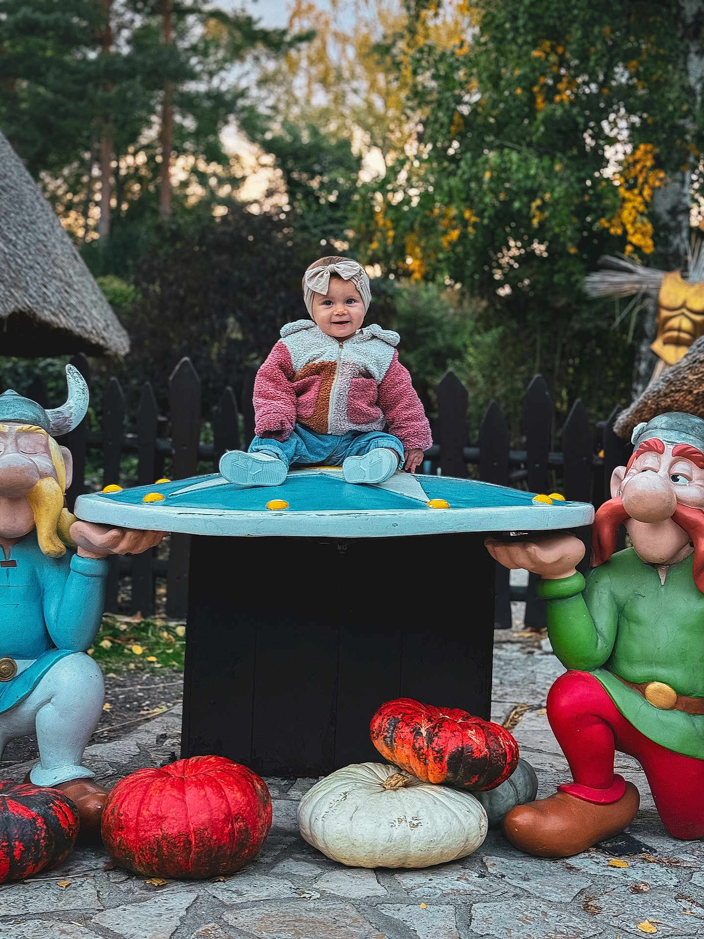 Bahia a rejoint le concours — aidez-le/la à gagner de superbes lots ! baby, child, outdoor, autumn, pumpkin, statue, viking, table, colorful_clothing, headband, smiling, nature, trees, fence, cobblestone, playful, seasonal, decoration, park, daylight