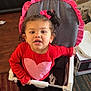 toddler, child, high_chair, red_shirt, pink_heart, curly_hair, bow, indoor, carpet, furniture, face, expression, socks, black_pants, toy_or_box, floor, person, seat, home, cute