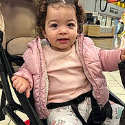 Neïla a rejoint le concours — aidez-le/la à gagner de superbes lots ! child, toddler, stroller, pink_jacket, curly_hair, shopping_mall, indoor, seat, harness, cute, smile, baby, clothing, portrait, eyes, hand, floor_tiles, expression, coat, pants
