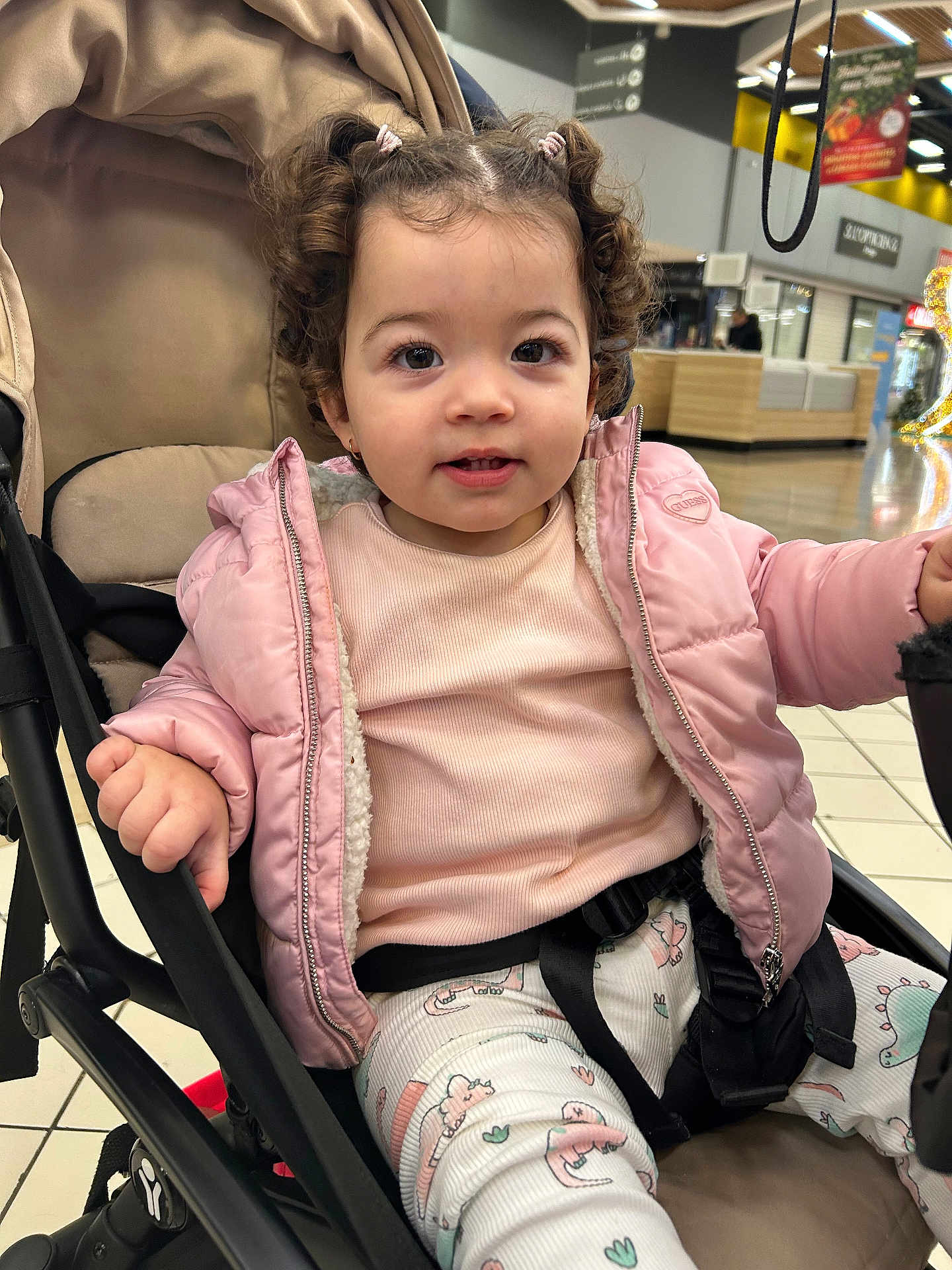 Neïla a rejoint le concours — aidez-le/la à gagner de superbes lots ! child, toddler, stroller, pink_jacket, curly_hair, shopping_mall, indoor, seat, harness, cute, smile, baby, clothing, portrait, eyes, hand, floor_tiles, expression, coat, pants