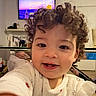 arm, child, clothing, curly_hair, cute, face, furniture, glass_table, indoors, living_room, pajamas, portrait, selfie, shelf, smile, teeth, television, toddler, toy, tv_screen