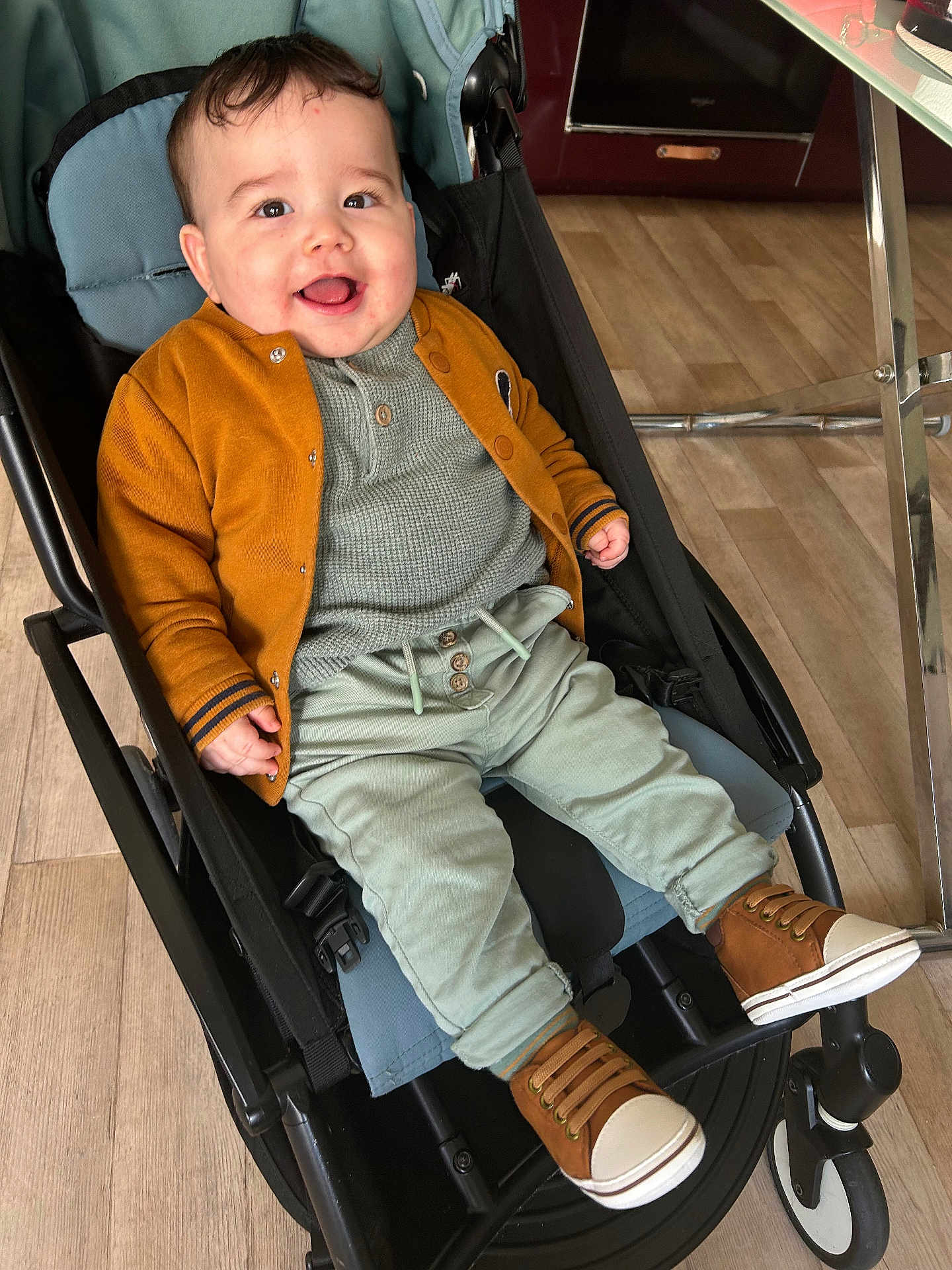Ishaq a rejoint le concours — aidez-le/la à gagner de superbes lots ! baby, child, stroller, smile, happy, jacket, cardigan, knit_top, green_pants, brown_shoes, sneakers, seat, indoor, wood_floor, metal_table, hand, face, eyes, cheeks, portrait