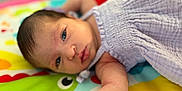 Eliana is registered to the contest to win money with this photo: baby, infant, face, arm, purple_clothing, play_mat, colorful, lying_down, skin, eyes, head, newborn, soft_texture, cute, person, child, young, indoors, closeup, expression