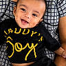 baby, child, smile, happy, face, person, clothing, black_shirt, yellow_text, sitting, lap, adult_arm, patterned_clothing, portrait, indoor, cute, joy, young_child, head, skin