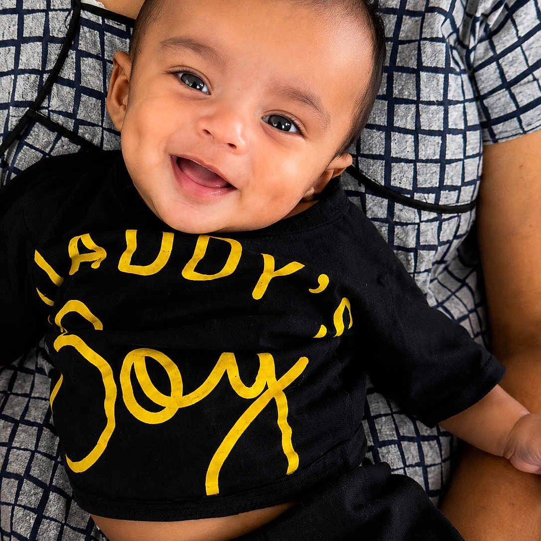 Niyansh joined the competition — help win amazing prizes! baby, child, smile, happy, face, person, clothing, black_shirt, yellow_text, sitting, lap, adult_arm, patterned_clothing, portrait, indoor, cute, joy, young_child, head, skin