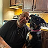 appliance, beard, black_shirt, bracelet, cabinet, collar, countertop, dog, expression, indoor, jewelry, kitchen, man, pet, playful, portrait, stove, tattoo, wooden_cabinet, yellow_wall