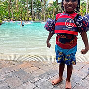 Davion is registered to the contest to win money with this photo: child, smiling, pool, swim_vest, swim_trunks, barefoot, water, outdoor, tropical, palm_trees, playful, happy, summer, vacation, people, recreation, sunlight, fun, holiday, posing