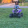 child, smiling, toy_atv, grass, outdoor, braided_hair, purple, vehicle, lawn, suburban, car, house, happy, play, daylight, person, nature, recreation, kid, fun