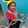 Xiomara is registered to the contest to win money with this photo: child, girl, smiling, hard_hat, safety_vest, control_panel, buttons, indoor, playful, short_sleeves, black_skirt, medical_tube, chair, carpet, person, bright_colors, toy, technology, happy, interactive