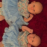 Evangeline And Anna is registered to the contest to win money with this photo: baby, twin, infant, blue_tutu, floral_clothing, lying_down, red_background, cute, small_hands, face, person, newborn, adorable, soft_texture, indoor, portrait, matching_outfits, child, smiling_eyes, young