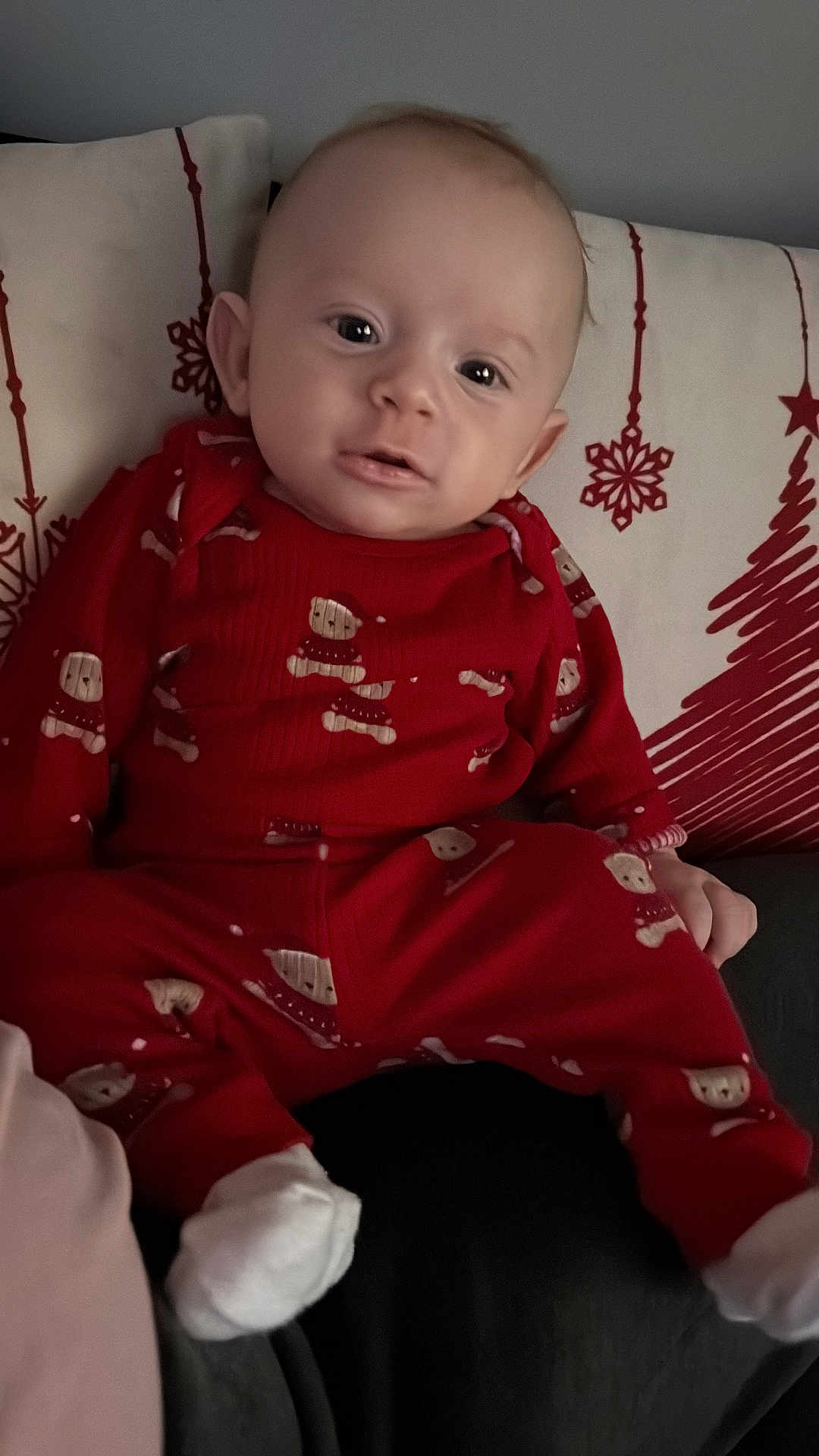 Liridon joined the competition — help win amazing prizes! baby, infant, child, red_pajamas, holiday_pattern, pillow, cushion, couch, sitting, indoor, portrait, face, eyes, ears, socks, feet, hands, cozy, adorable, neutral_expression