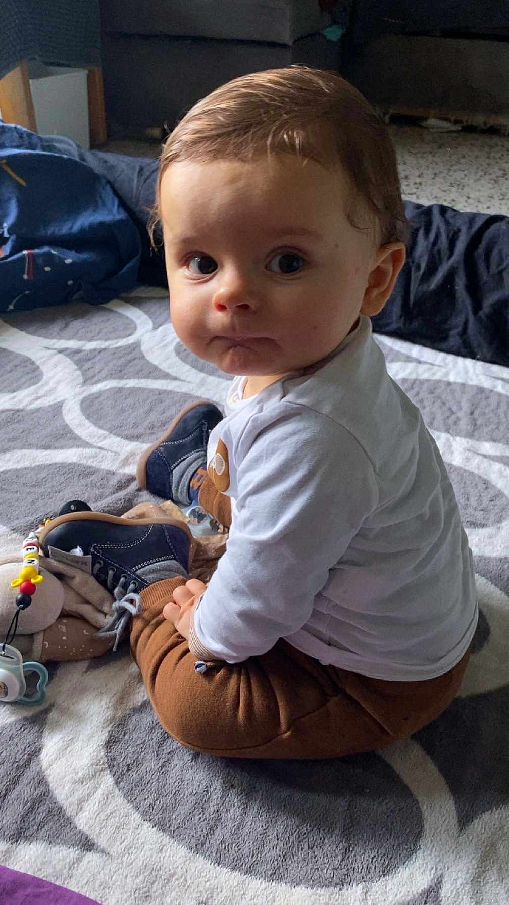 Adrien participe au concours pour gagner de l'argent avec cette photo : baby, child, sitting, carpet, toy, shoe, clothing, indoors, floor, curious, face, person, footwear, white_shirt, brown_pants, pacifier, soft_toy, patterned_rug, home, young