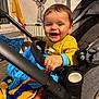 baby, child, smiling, stroller, blanket, indoor, yellow_clothing, hand, face, happy, toy, window, furniture, decorations, bat_decor, blue_blanket, orange_blanket, person, cozy, portrait