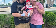 Nova is registered to the contest to win money with this photo: family, baby, man, woman, outdoor, tree, grass, sky, clouds, smiling, casual_clothing, shorts, tshirt, footwear, headband, bib, celebration, holiday, summer, nature