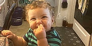 Antonio is registered to the contest to win money with this photo: child, toddler, curly_hair, striped_shirt, cartoon_leggings, kitchen, floor_mat, stove, refrigerator, washing_machine, laundry_basket, person, smile, nose_picking, indoor, cabinet, towel, chair, hand