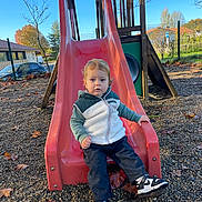Alessio participe au concours pour gagner de l'argent avec cette photo : child, toddler, slide, playground, outdoor, fall, autumn_leaves, pebbled_ground, casual_clothing, sneakers, hooded_jacket, curly_hair, daylight, trees, fence, residential_area, park, play_equipment, person, young_child