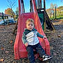 child, toddler, slide, playground, outdoor, fall, autumn_leaves, pebbled_ground, casual_clothing, sneakers, hooded_jacket, curly_hair, daylight, trees, fence, residential_area, park, play_equipment, person, young_child