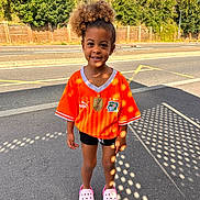 Elayana participe au concours pour gagner de l'argent avec cette photo : casual_clothing, child, curly_hair, daylight, fence, happy, orange_jersey, outdoor, person, pink_crocs, portrait, shadow_pattern, shorts, sidewalk, smiling, standing, street, sunlight, trees, young
