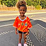 casual_clothing, child, curly_hair, daylight, fence, happy, orange_jersey, outdoor, person, pink_crocs, portrait, shadow_pattern, shorts, sidewalk, smiling, standing, street, sunlight, trees, young