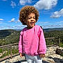 blue_sky, child, clouds, curly_hair, daylight, happy, hills, map_display, nature, outdoor, person, pink_sweater, portrait, railing, smiling, standing, stone_wall, sunny, toddler, white_pants