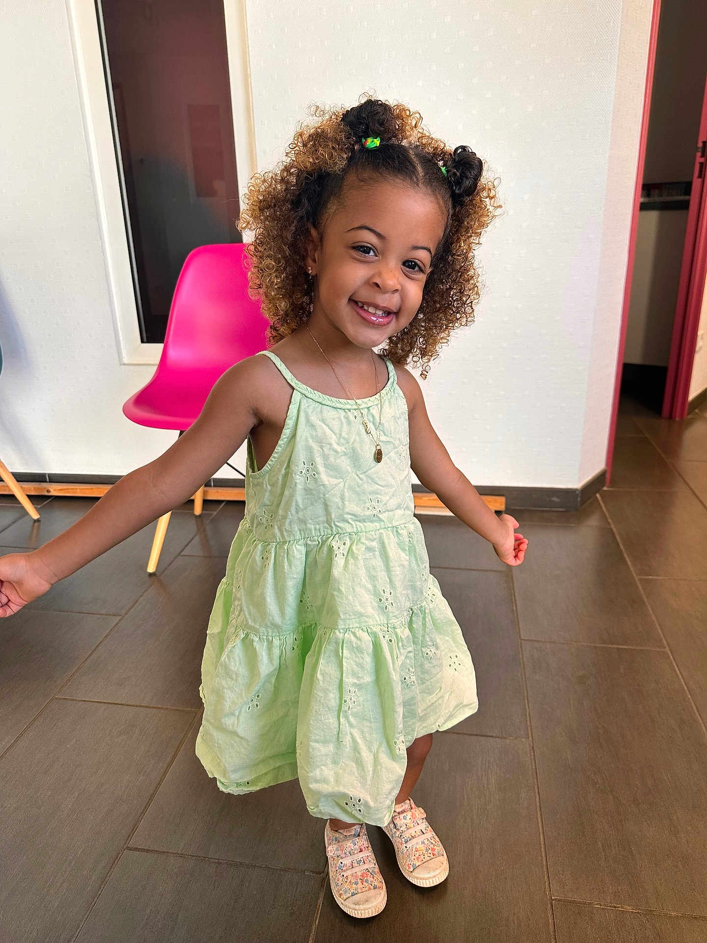Elayana participe au concours pour gagner de l'argent avec cette photo : chair, child, curly_hair, cute, dress, floor, floral_sneakers, girl, green_dress, happy, indoor, jewelry, necklace, person, pink_chair, playful, smiling, sneakers, standing, tile_floor