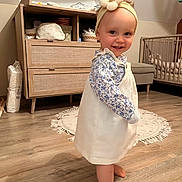 Mila a rejoint le concours — aidez-le/la à gagner de superbes lots ! toddler, child, barefoot, dress, headband, smile, indoor, wooden_floor, nursery, furniture, cabinet, crib, rug, baby, person, flooring, clothing, headwear, happy, home
