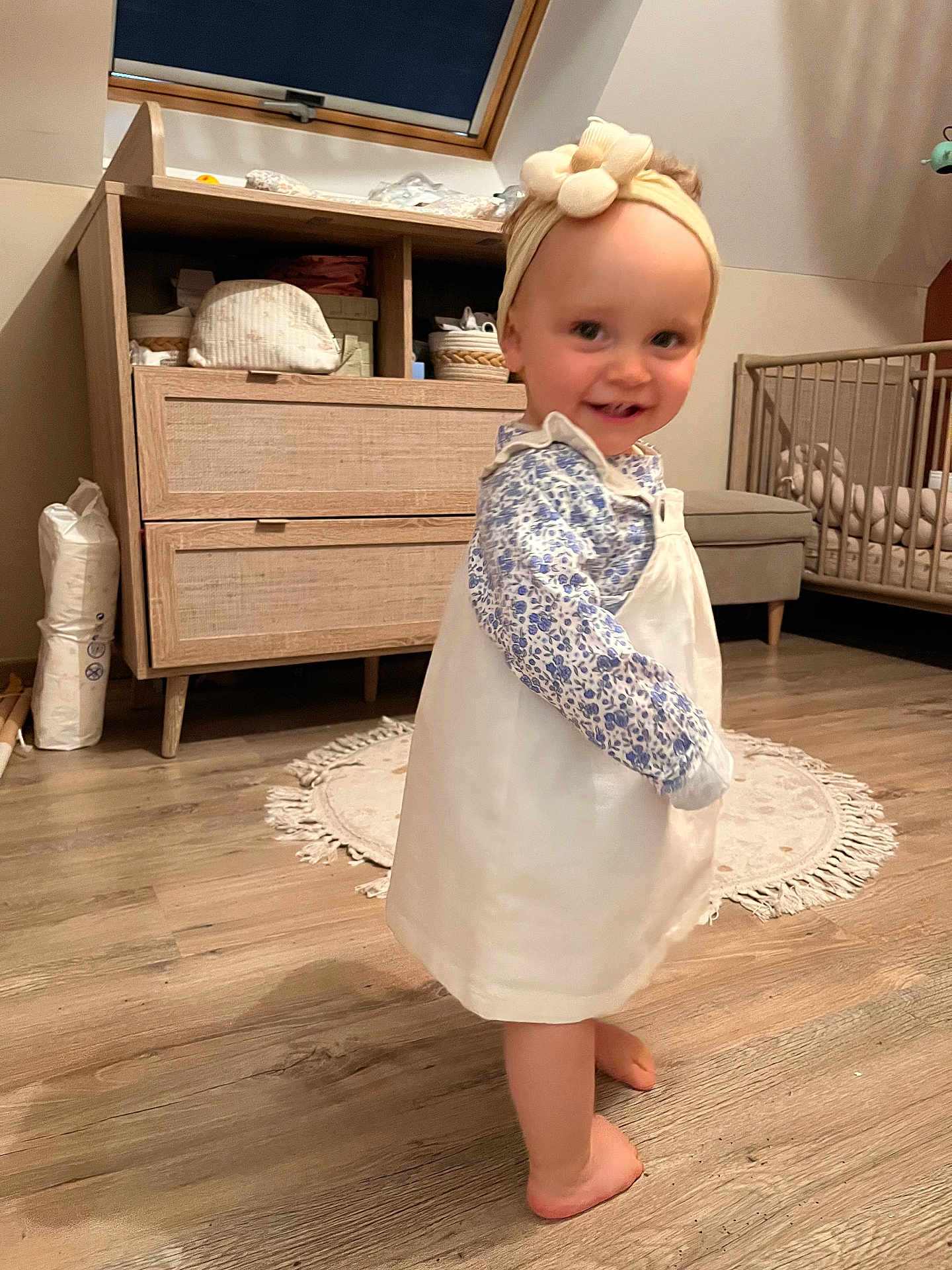 Mila a rejoint le concours — aidez-le/la à gagner de superbes lots ! toddler, child, barefoot, dress, headband, smile, indoor, wooden_floor, nursery, furniture, cabinet, crib, rug, baby, person, flooring, clothing, headwear, happy, home