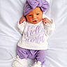 baby, newborn, infant, purple_bow, white_sweater, purple_pants, white_booties, lying_down, blanket, cute, portrait, child, soft_texture, cozy_clothing, small_hands, face, headwear, smiling, indoors, sleepwear