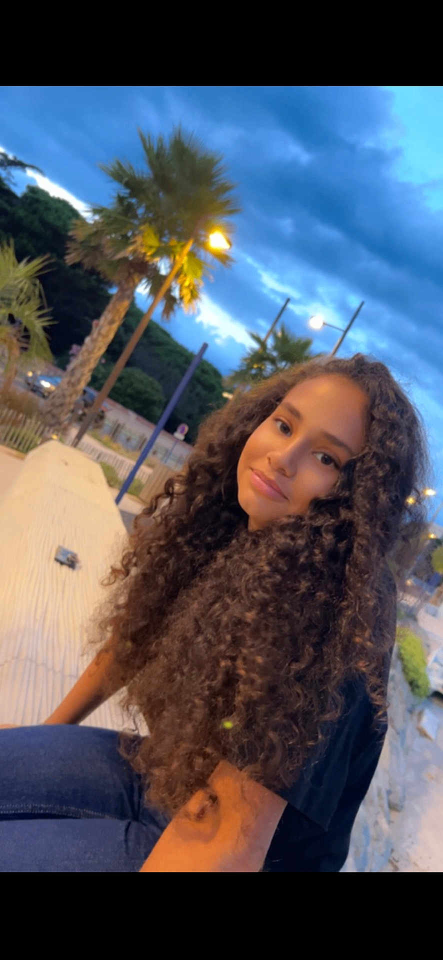 Ilhem participe au concours pour gagner de l'argent avec cette photo : girl, curly_hair, outdoor, twilight, palm_tree, street_lamp, smile, jeans, black_shirt, cloudy_sky, nature, park, bench, young_person, portrait, happy, casual, evening, scenery, relaxation