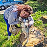 woman, dog, tree_stump, grass, parked_cars, outdoor, leisure, casual_clothing, glasses, sunny_day, pet, animal, nature, smiling, blue_eyes, leash, pavement, urban, friendship, daylight
