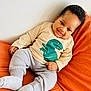 Kayce is registered to the contest to win money with this photo: baby, child, smiling, dinosaur_shirt, beige_shirt, gray_pants, socks, orange_cushion, indoor, portrait, cute, happy, infant, relaxed, seated, cozy, young_child, face, person, casual_clothing