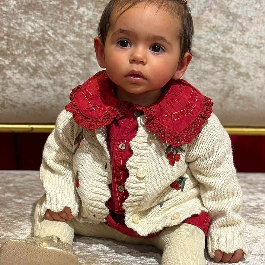 Sevgi participe au concours pour gagner de l'argent avec cette photo : baby, cardigan, child, closeup, curious_expression, cute, face, footwear, head, indoor, knitwear, person, pigtails, plush_surface, portrait, red_clothing, sitting, soft_texture, toddler, warm_clothing