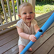 Claire is registered to the contest to win money with this photo: baby, child, smiling, blue_pole, wooden_floor, porch, outdoor, sunlight, greenery, railing, playful, happy, infant, person, short_sleeves, barefoot, necklace, daylight, cute, closeup