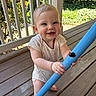 baby, child, smiling, blue_pole, wooden_floor, porch, outdoor, sunlight, greenery, railing, playful, happy, infant, person, short_sleeves, barefoot, necklace, daylight, cute, closeup