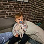 toddler, child, pacifier, stuffed_animal, pigtails, couch, pillow, blanket, brick_wall, tablet, charging_cable, indoor, casual_clothing, long_sleeve_shirt, pants, socks, cute, seated, toy, home