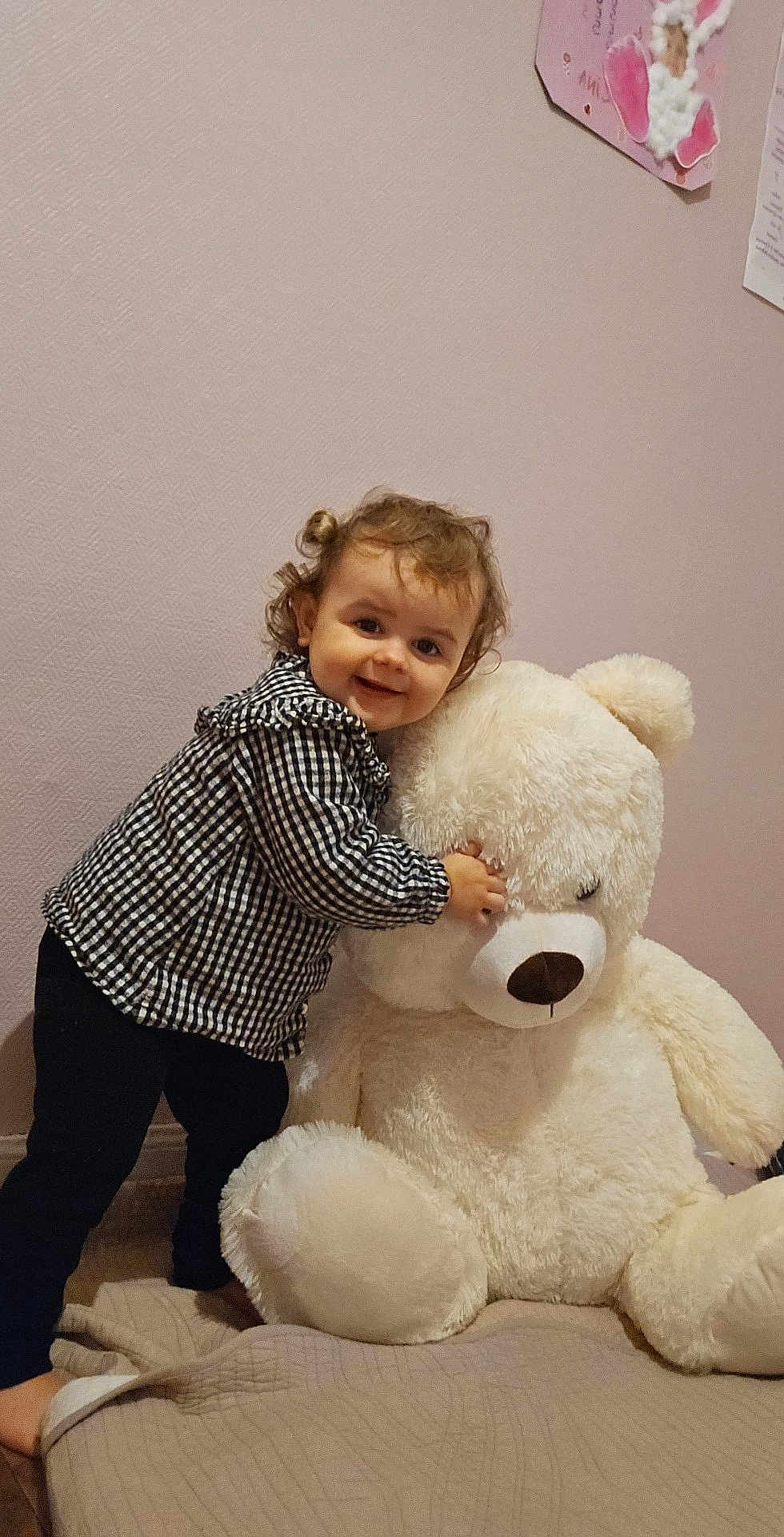 Lina a rejoint le concours — aidez-le/la à gagner de superbes lots ! toddler, child, smile, curly_hair, teddy_bear, stuffed_animal, bed, blanket, indoor, wall, artwork, toy, happy, person, cuddly, plush, home, cozy, cute, black_pants