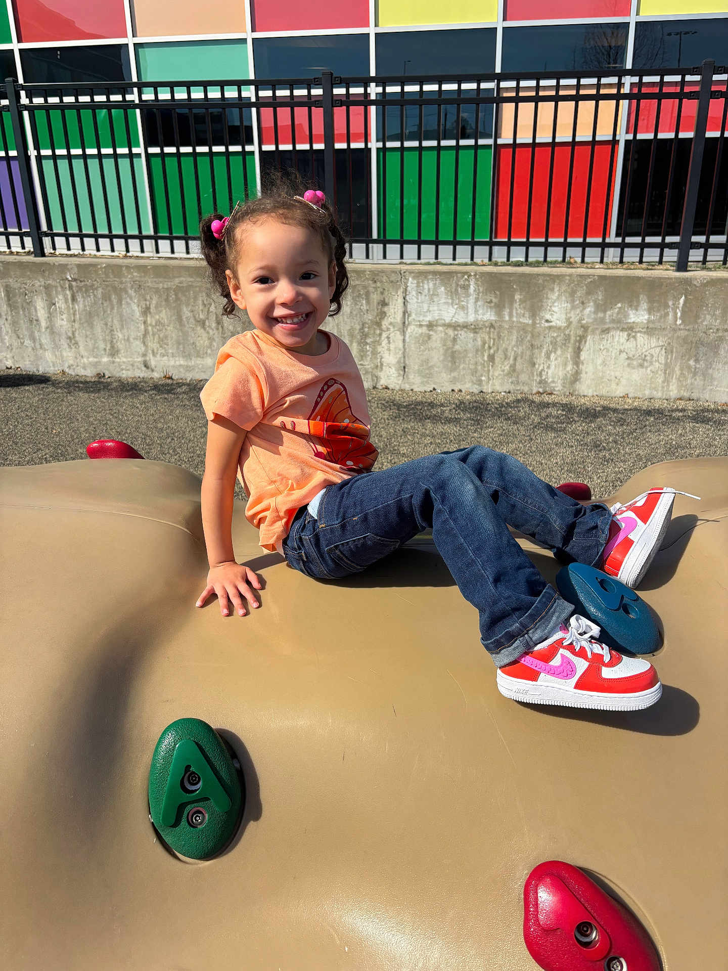 Harmony joined the competition — help win amazing prizes! child, smiling, playground, climbing_structure, sneakers, pink_hair_ties, orange_shirt, jeans, outdoor, sunny, fence, colorful_wall, concrete, happy, person, young, casual_clothing, daylight, recreation, play