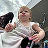 toddler, child, stroller, blanket, hand, face, person, outdoor, cloudy_sky, building, pouting, beads, clothing, adult, background, urban, expression, portrait, baby, people