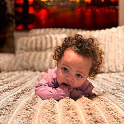 Amani joined the competition — help win amazing prizes! baby, infant, child, curly_hair, blue_eyes, teething, hand_in_mouth, pink_clothes, blanket, bed, lying_down, cozy, soft_texture, bokeh, portrait, indoor, home, background_painting, cute, adorable
