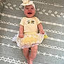 baby, smiling, child, yellow_outfit, headband, bow, lying_down, patterned_mat, happy, cute, infant, clothing, indoors, toddler, legs, hands, face, baby_girl, white_lace, joy
