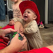 Greyson joined the competition — help win amazing prizes! baby, child, red_hat, smiling, video_game_controller, adult_arm, indoor, flooring, door, fan, toy, blurred_child, happy, casual_clothing, home, person, playing, candid, joyful, interaction